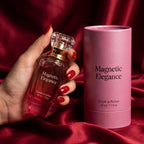 Hand with red manicure holding the Magnetic Elegance Extrait de Parfum bottle on red silk beside its presentation box